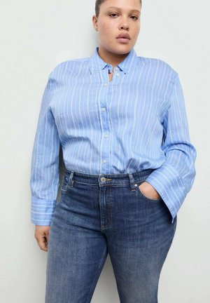 Person trägt ein blau-weiß gestreiftes Hemd mit Knöpfen, das in eine hoch taillierte dunkelblaue Jeans gesteckt ist, eine Hand in der Tasche, neutraler Hintergrund.