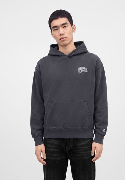 Νεαρός άνδρας που φοράει σκούρο γκρι hoodie Billionaire Boys Club με μπροστινή τσέπη και μαύρα τζιν μπροστά σε ένα απλό ανοιχτό φόντο.