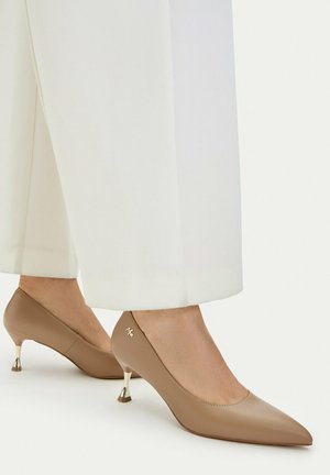 Jasnobrązowe skórzane czółenka kitten heels z zaostrzonym noskiem i złoconym, zakrzywionym obcasem, noszone z białymi spodniami kończącymi się powyżej kostki.