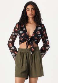 Blouse noire transparente avec des motifs de coquillages colorés et un devant noué ; associée à un short léger vert olive avec un waistband élastique.