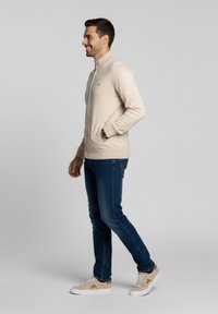 Chaqueta de punto beige con cremallera y bolsillos laterales, llevada sobre unos vaqueros azul oscuro y zapatillas beige. La modelo está de perfil frente a un fondo gris.