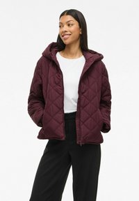 Maroon quilted jacket with a hood, featuring diagonal stitching, a zipper front, and side pockets, worn over a white top and black pants.