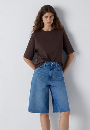 Young woman with long curly hair wearing a loose dark brown t-shirt and knee-length blue denim shorts standing against a light wall.