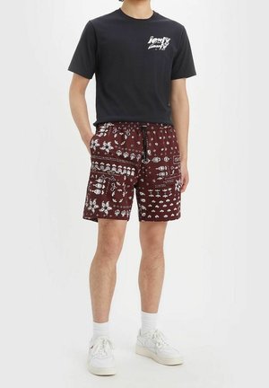 Bordeauxfarbene Shorts mit weißen Musterdetails, elastischem Bund und Kordelzug. Kombiniert mit einem schwarzen T-Shirt mit einem Logo. Weiße Sneakers.
