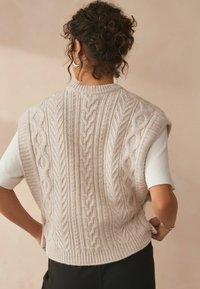 Neule sleeveless -neule vaaleanbeigessä, jossa on kierrätysmateriaaleilla virkattuja kuvioita, ribattu helma ja pyöreä pääntie, käytetään lyhythihaisen valkoisen paidan päällä.