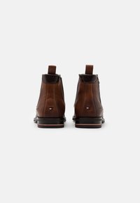 Botas de cuero marrón hasta el tobillo con un acabado suave, lengüetas de tiro, tacones bajos y un pequeño detalle de logo en el lado exterior.