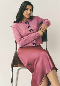 Rosa stickad tröja med svarta rosettaccent, kombinerad med en glänsande rosa satinkjol, sittande på en metallstol mot en neutral bakgrund.