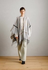 Poncho grigio in lana con bordi frangiati, caratterizzato da un motivo a righe in crema e marrone, indossato sopra una maglietta chiara e pantaloni crema.
