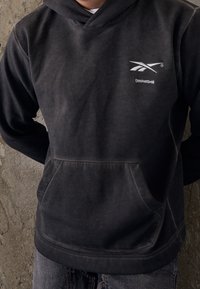 Felpa con cappuccio grigio scuro, dotata di tasca frontale, con un logo bianco sul petto. Il tessuto ha un aspetto liscio e testurizzato.