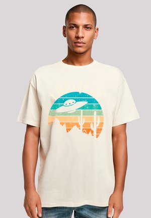 Beige Baumwoll-T-Shirt mit einem grafischen Design, das ein Alien-Raumschiff, Berge und Kakteen in Farbverläufen von Grün, Orange und Blau zeigt.