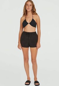 Mlada žena stoji, odjevena u crni halter bikini top, crne šortse s vezicom i crne sandale na otvorenom petu, na jednobojnoj pozadini.