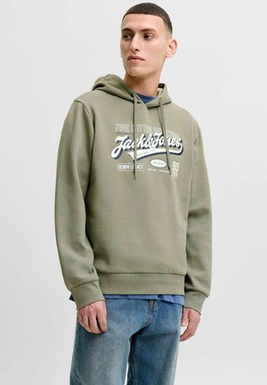 Ung mand iført en olivengrøn Jack & Jones hættetrøje og cowboybukser, der kigger let til venstre mod en ensartet lys baggrund.