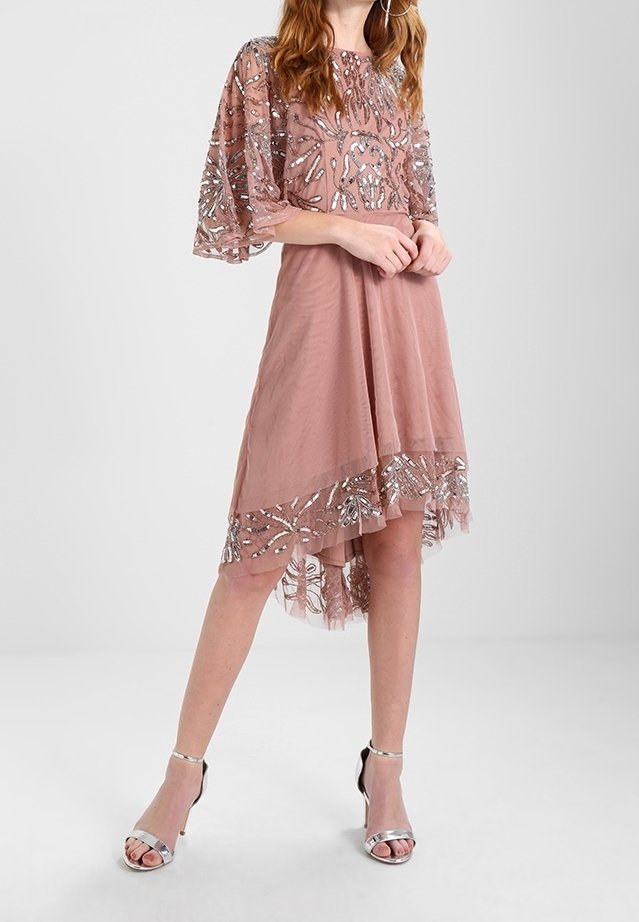 Femme portant une robe rose poudré avec des motifs floraux pailletés, des manches transparentes jusqu'au coude et des sandales argentées à talons hauts, se tenant debout les mains jointes.
