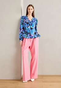 Blusa floreale con uno sfondo nero decorata con fiori blu, rosa e verdi, abbinata a pantaloni lunghi rosa e scarpe bianche a punta.