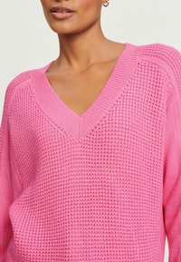 Frau in einem pinken Waffelstrick-V-Ausschnitt-Pullover mit geripptem Kragen und Schulternahtdetails, dargestellt von Hals bis zur Mitte des Oberkörpers.