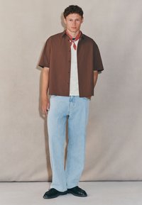 Brown short-sleeve button-up shirt with black buttons, white t-shirt underneath, light blue straight-leg jeans, and black shoes. Red bandana tied around the neck.