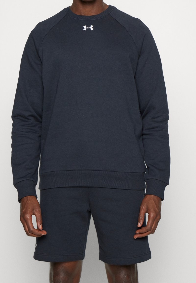 Uomo che indossa una felpa nera a maniche lunghe di Under Armour e pantaloni corti neri abbinati, in piedi su uno sfondo bianco uniforme.