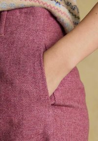 Różowe spodnie w jodełkę z boczną kieszenią. Materiał wydaje się mieć fakturę, ukazując subtelny wzór. Częściowo widoczna ręka.