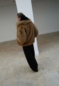 Giacca di pelliccia sintetica marrone con colletto ampio, vestibilità oversize e strato sottostante con cappuccio nero. Abbinata a pantaloni neri larghi.