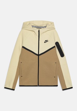 Jachetă cu glugă Nike, cu panouri bej și maro, accente negre, închidere cu fermoar și un mic buzunar cu fermoar pe mânecă.