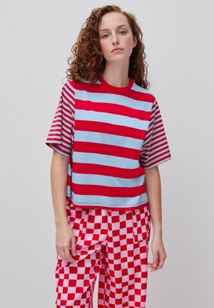 Jonge vrouw met krullend rood haar die een rood-lichtblauw gestreept shirt en rood-roze geruite broek draagt, staand voor een effen achtergrond.