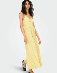 Robe maxi jaune à bretelles spaghetti, avec un bodice noué et une jupe à volants. Le tissu a une texture douce, complétant un style décontracté.
