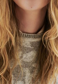 Pull tricot�é marron clair avec un motif texturé, doté d'un col côtelé. Cheveux longs, ondulés et clairs partiellement visibles.