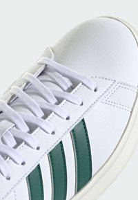 Biele kožené tenisky s bielymi šnúrkami, ktoré majú na boku tri zelené pruhy a textúrovaný povrch. Viditeľná gumová podrážka.