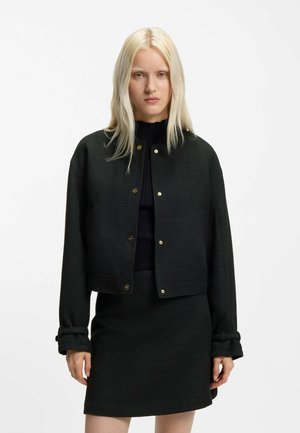 Frau trägt eine schwarze, geknöpfte Jacke über einem schwarzen Oberteil und einem passenden schwarzen Rock, steht vor einem schlichten weißen Hintergrund.