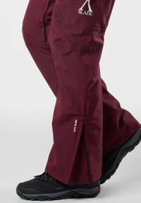 Burgundowe wodoodporne spodnie o eleganckim kroju, z bocznymi zamkami błyskawicznymi i białym logo. W zestawieniu z czarnymi, wytrzymałymi butami. Tekstura wydaje się gładka i trwała.