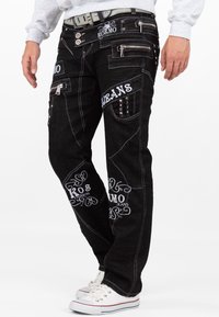 Jeans neri con cuciture bianche, numerose tasche e ricami decorativi bianchi. Presentano cerniere e borchie sulle tasche.