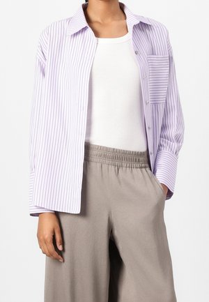 Persoon draagt een wit tanktop, lavendel- en witgestreept overhemd met knopen en beige broek met elastische taille, met één hand in de zak.