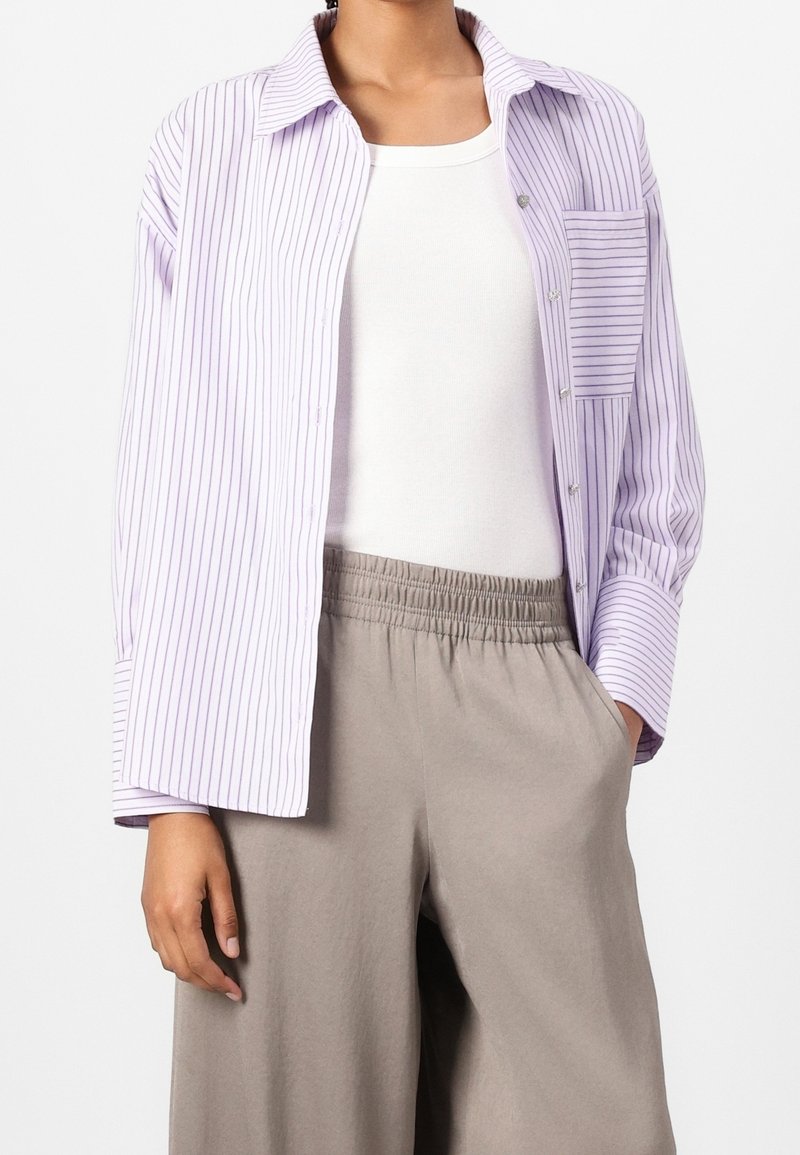 Personne portant un débardeur blanc, une chemise rayée lavande et blanche à boutons, et un pantalon beige taille élastique, une main dans la poche.