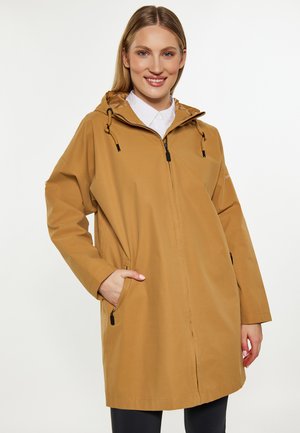 Eine lächelnde Frau, die eine beige Kapuzenjacke mit Reißverschluss und Taschen sowie ein weißes Hemd darunter trägt, vor einem schlichten weißen Hintergrund.