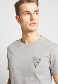 Camiseta de algodón gris con mangas cortas que presenta un diseño gráfico negro de montañas y un sol dentro de un triángulo en la zona izquierda del pecho.