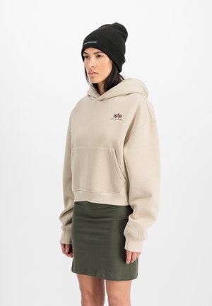 Beige cropped hoodie met voorzak en logo; gecombineerd met een donkergroene getailleerde rok. Een zwarte geribbelde beanie maakt de look af.