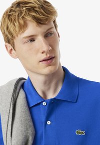 Nuori mies, jolla on vaaleat hiukset, on pukeutunut siniseen polo-paitaan ja katsoo hiukan sivulle. Harmaa villatakki on heitetty hänen olkapäälleen.