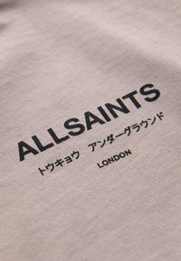 Béžová tkanina s čiernym textom "ALLSAINTS" a japonskými znakmi nad slovom "LONDON" potlačená na nej.