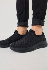 Czarne dzianinowe sneakersy z teksturowanym wzorem, bez zapięcia, na gumowej podeszwie. Noszone z podwiniętymi czarnymi dżinsami.