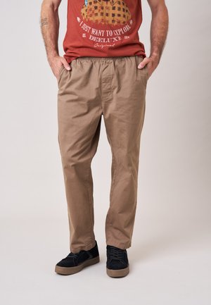 Man draagt lichtbruine casual broek met elastische taille en zwarte sneakers, staat met handen in de zakken tegen een effen achtergrond.