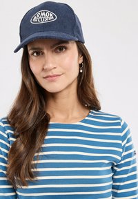 Cappello di velluto a coste blu navy con logo "ARMOR LUX" ricamato in bianco, indossato con una maglietta a righe blu e bianche. Lunghi capelli castani parzialmente visibili.