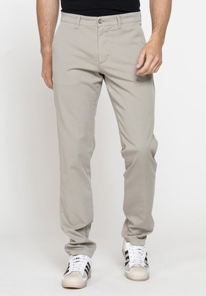 Uomo che indossa pantaloni casual beige chiaro e scarpe da ginnastica bianche con strisce nere, in piedi davanti a uno sfondo bianco uniforme.