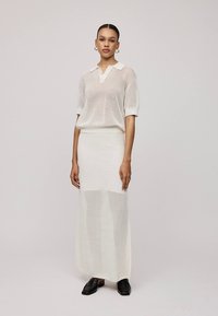 Hvit to-delt strikket antrekk med kortermet genser med krage og et langt, tettsittende skjørt med mønsterstruktur. Sorte sko.