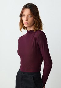 Burgunderfarbenes Langarmoberteil mit hohem Kragen, aus einem glatten, dehnbaren Stoff gefertigt. Figurbetontes Design; kombiniert mit schwarzen, hochgeschnittenen Hosen.