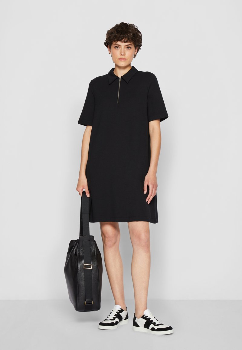 Svart kortärmad klänning med dragkedja vid kragen, tillverkad av mjukt material. Kombinerad med en stor svart tygväska och svart-vita sneakers.