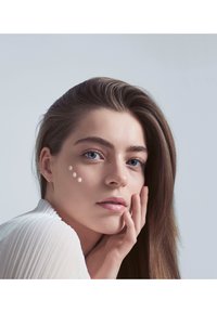 Close-up of a person with smooth skin, three cream dots on the cheek, long brown hair, wearing a textured white top against a light background.
