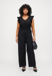 Black jumpsuit with ruffled sleeves, button detail on top, wide-legged pants, belted waist, paired with black heels and a clutch bag.