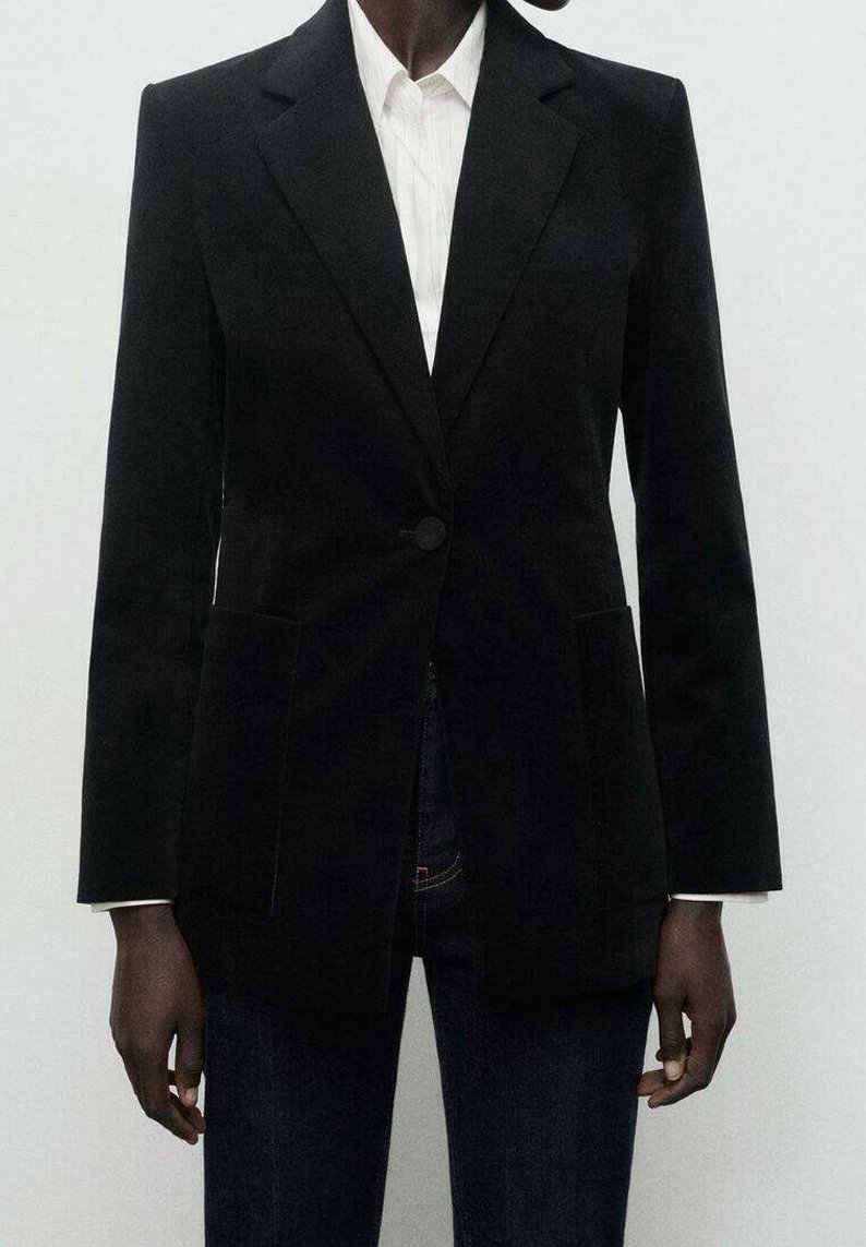 Personne portant un blazer noir sur mesure par-dessus une chemise blanche à col et un jean foncé, debout devant un fond uni.