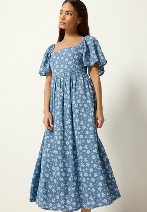 Joven mujer con un vestido largo azul con estampado floral y mangas abullonadas, mirando hacia un lado sobre un fondo claro y liso.