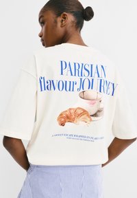 Krémovo sfarbené oversized tričko so potlačou croissantu a šálky, s modrým textom "PARÍZSKA chuťOVÁ CESTA."